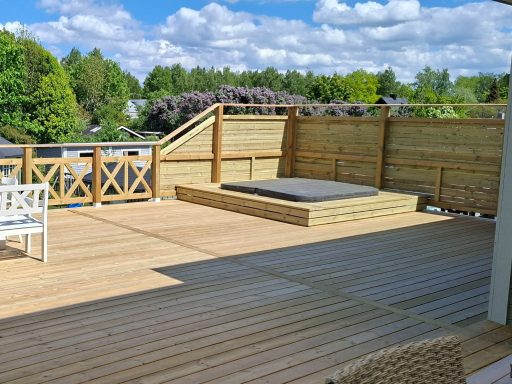Altanbygge med nedsänkt SPA bad Träaltan med skyddsräcke och ett platt område, omgiven av grönska och blå himmel.